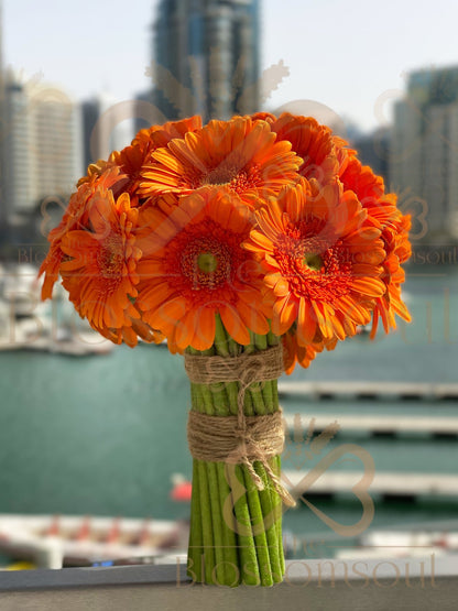 Orange gerberas bouquet tied with natural jute twine, standing upright against a blurred Dubai Marina skyline. Radiant floral design in warm direct sunlight.